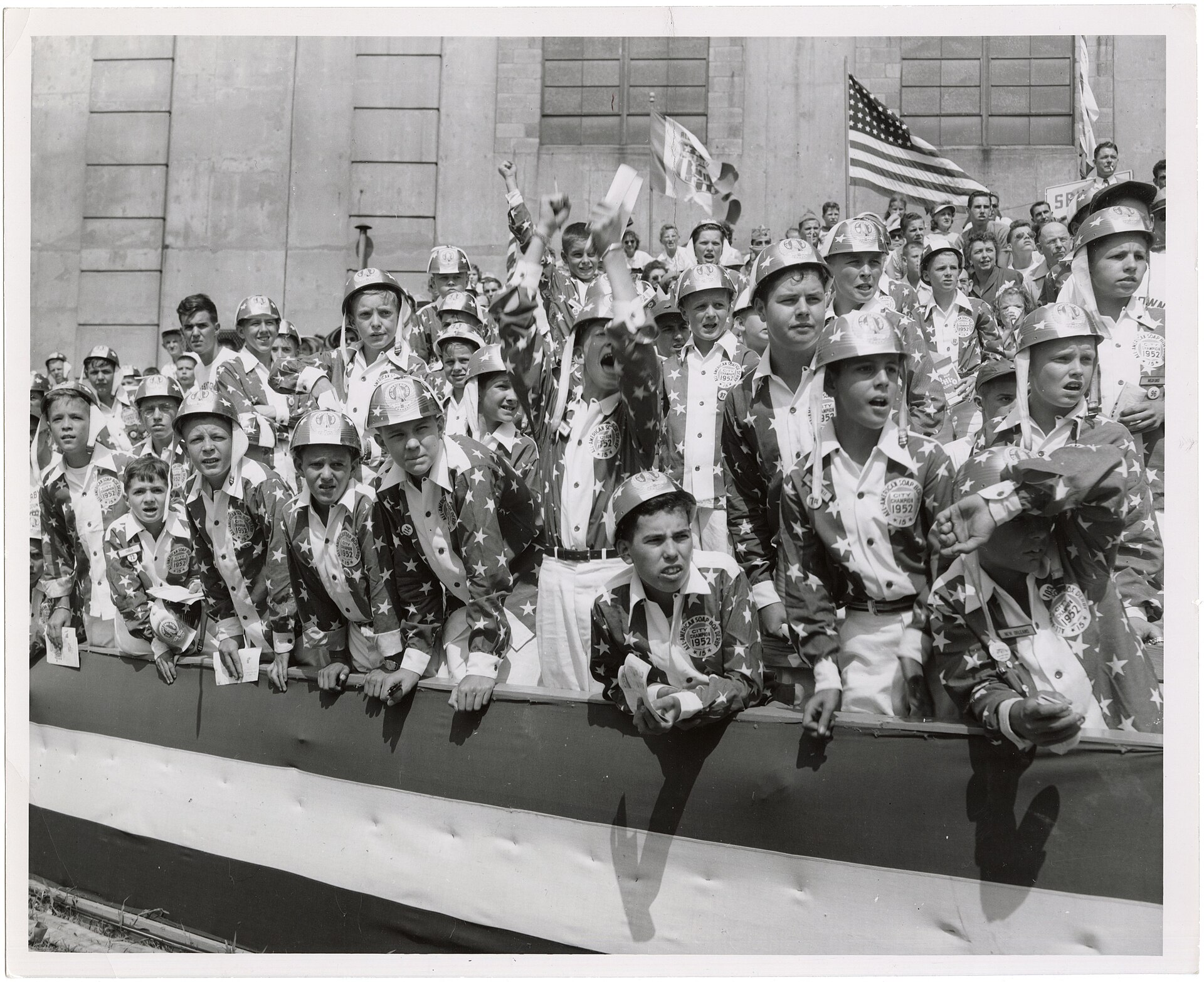 Young 1952 Soap Box Derby city champions in star-pattern uniforms cheering from a crowded railing outside Akron's Rubber Bowl