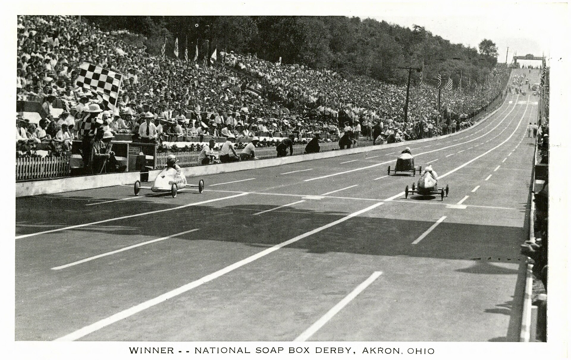 Three early Soap Box Derby cars cross the Derby Downs finish while a checkered flag waves and spectators cover both hillsides