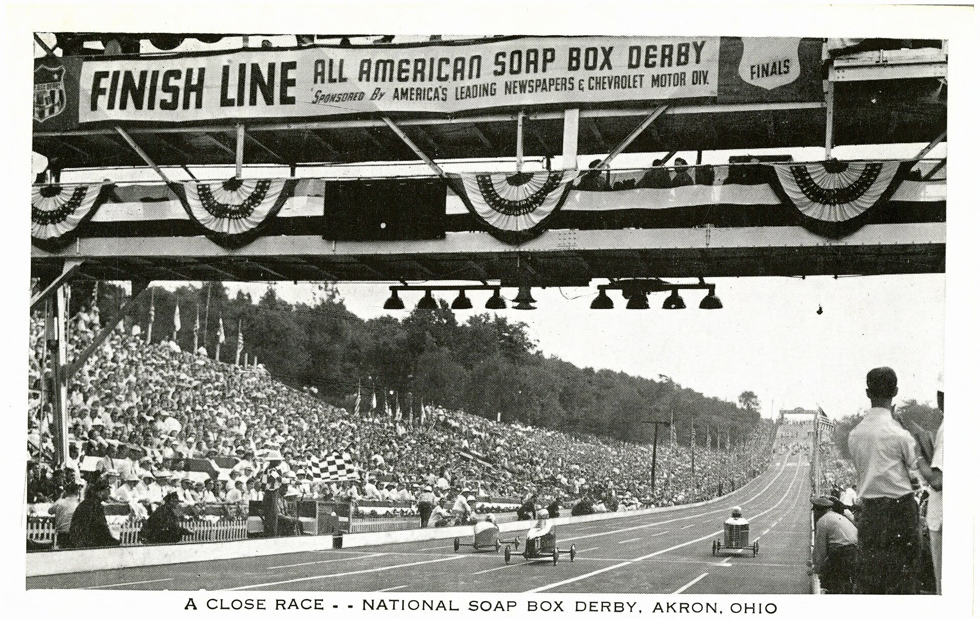 Three early Soap Box Derby cars racing toward the All-American finish structure as a packed hillside watches