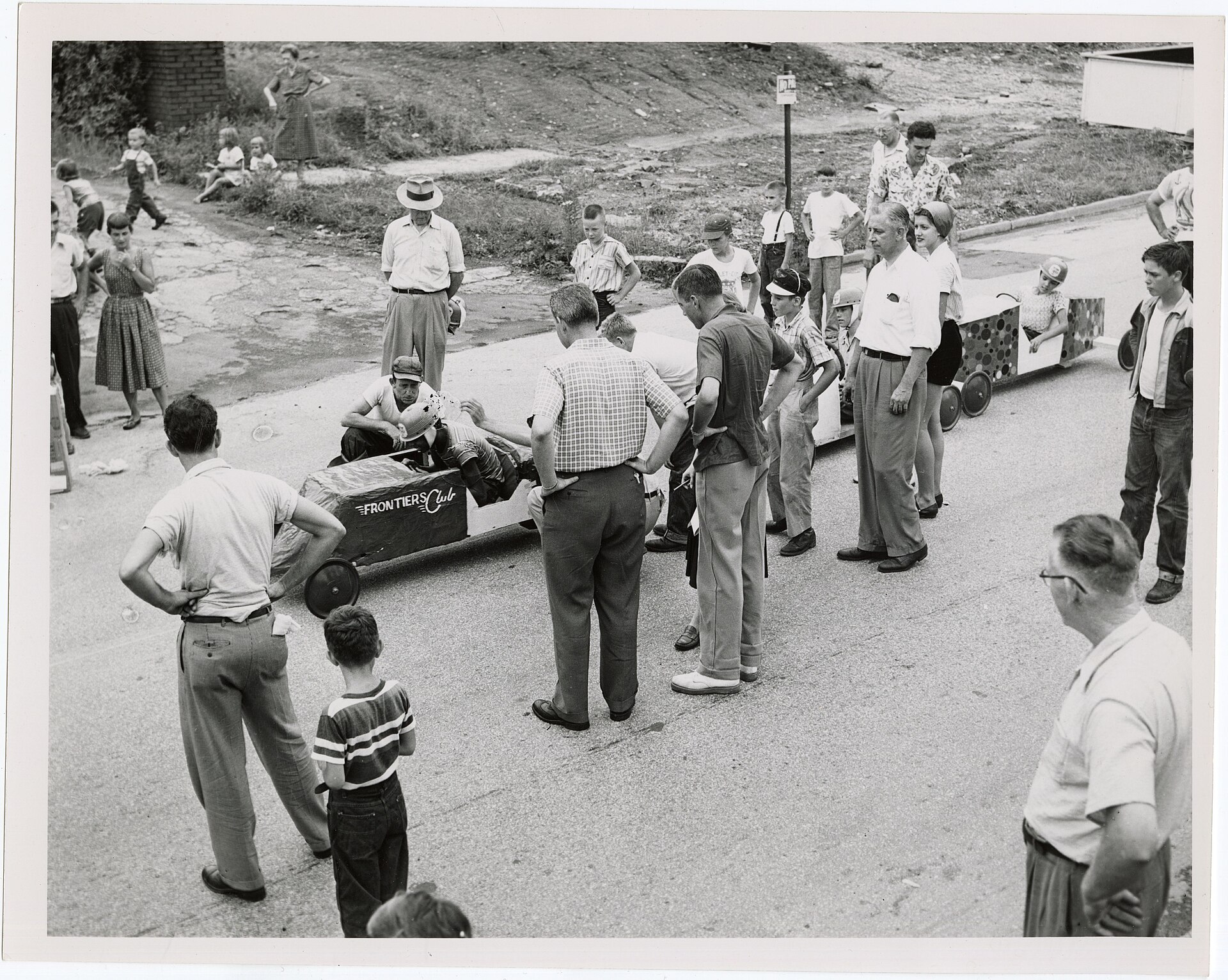 Three young racers seated in handmade Soap Box Derby cars at a neighborhood start line while families and officials watch