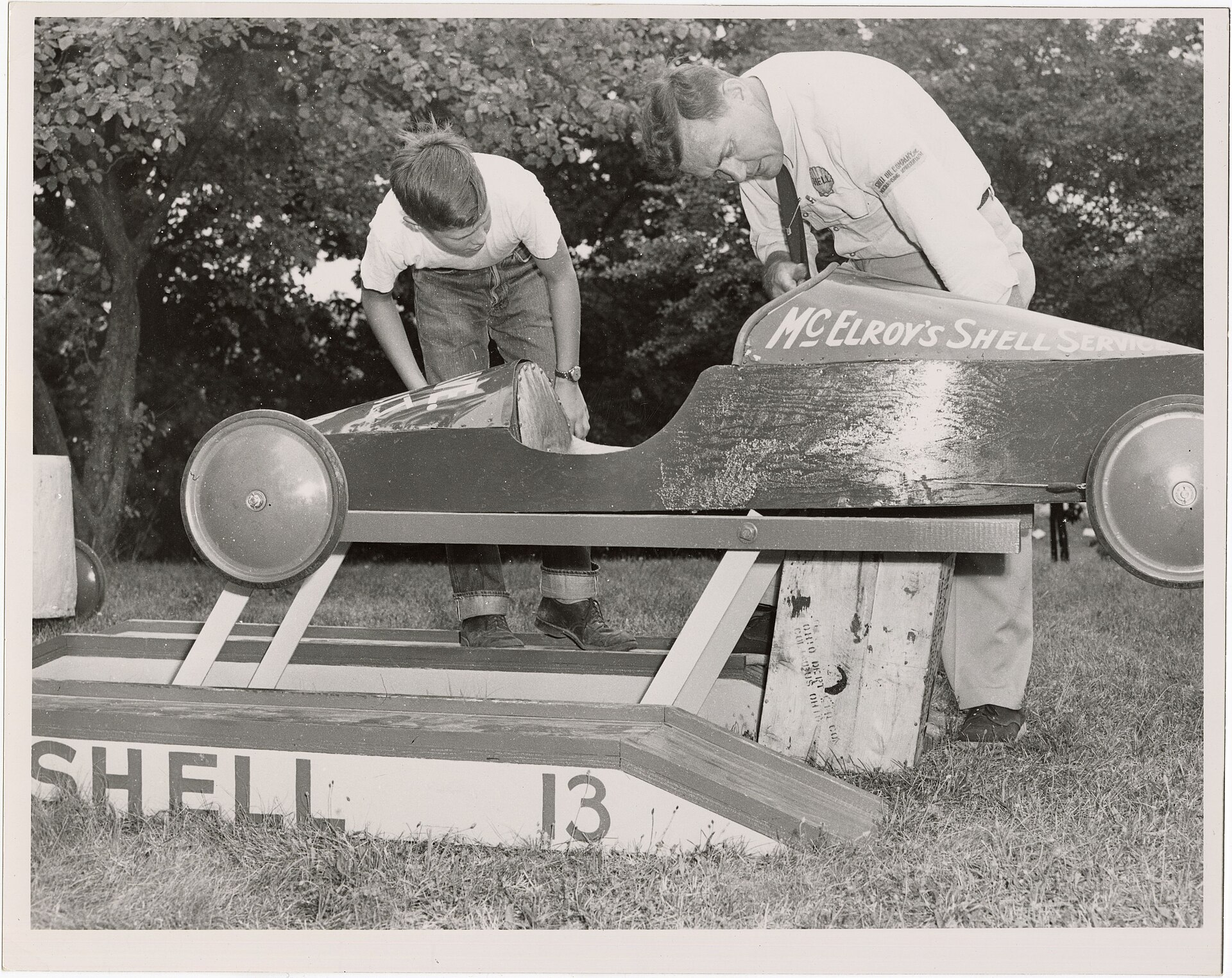 A young racer and a Shell representative inspect a long wooden Soap Box Derby car on a raised stand in 1953